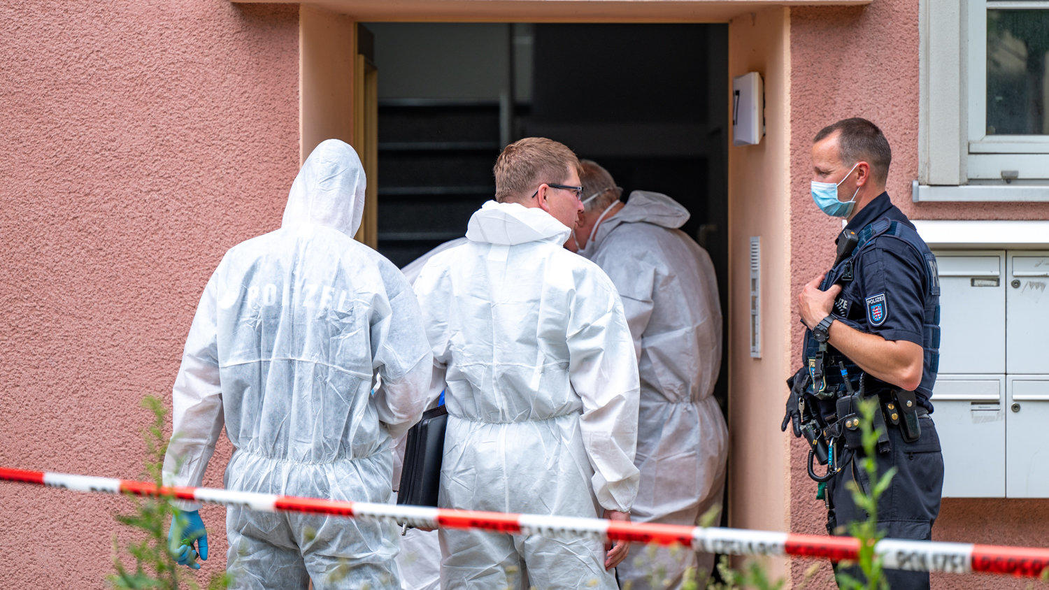 Kriminalbeamte stehen vor einem Haus, das mit rot-weißem Flatterband abgesperrt ist. In dem Gebäude wurde eine männliche Leiche gefunden. Die Ermittler gehen von einem Tötungsdelikt aus.