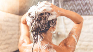 Back view of beautiful naked young woman is using shampoo while taking shower in bathroom
