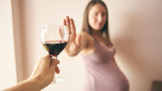 Eine schwangere Frau, der ein Glas Rotwein hingehalten wird, lehnt dieses ab und sagt damit "Nein" zu Alkohol während der Schwangerschaft.