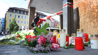 26.06.2021, Bayern, Würzburg: Ein Polizist steht neben Kerzen und Blumen vor einem geschlossenen und abgesperrten Geschäft in der Innenstadt. In Würzburg hat ein Mann am Vortag wahllos Menschen mit einem Messer attackiert. Drei Menschen starben, mindestens fünf wurden schwer verletzt. Foto: Karl-Josef Hildenbrand/dpa +++ dpa-Bildfunk +++