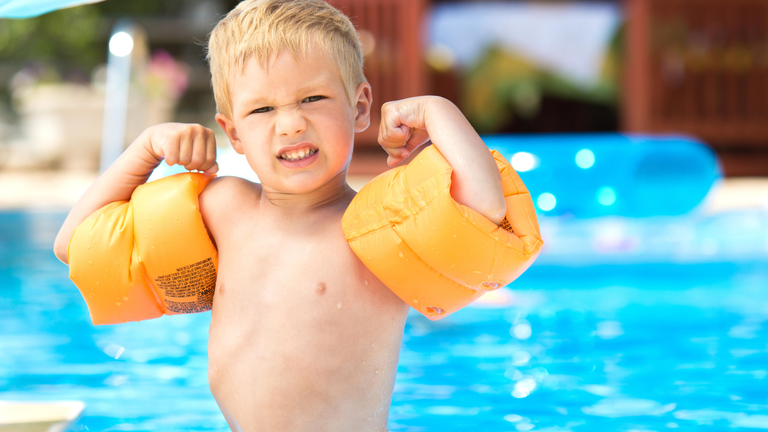 Ein kleiner Junge steht am Beckenrand eines Schwimmbads und zeigt seine starken Armmuckis. An seinen Armen trägt er Schwimmflügel.