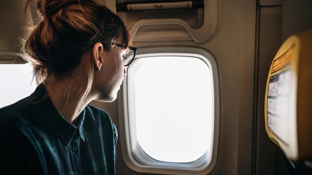 Eine junge Frau sitzt im Flugzeug auf einem Fensterplatz und schaut aus dem Fenster in den Himmel.