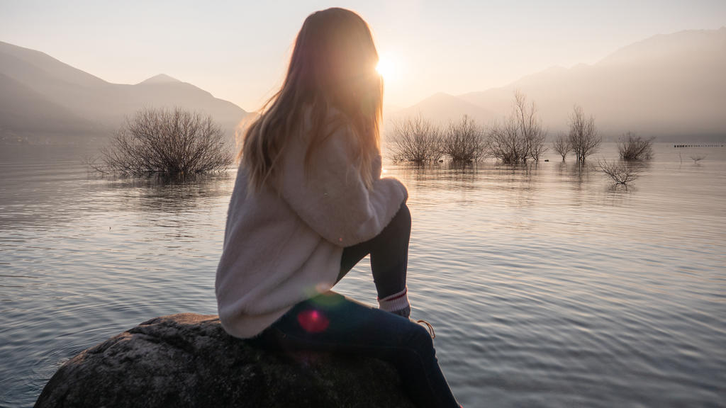 Eine junge Frau sitzt bei Sonnenuntergang auf einem Stein am See und schaut in die Ferne.