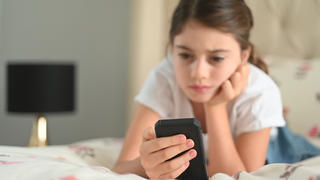 Ein junges Mädchen sitzt auf dem Bett und hält ihr Handy in der Hand. Ihrem Gesichtsausdruck nach scheint sie bedrückt und etwas gesehen zu haben, was ihr nicht gefällt.