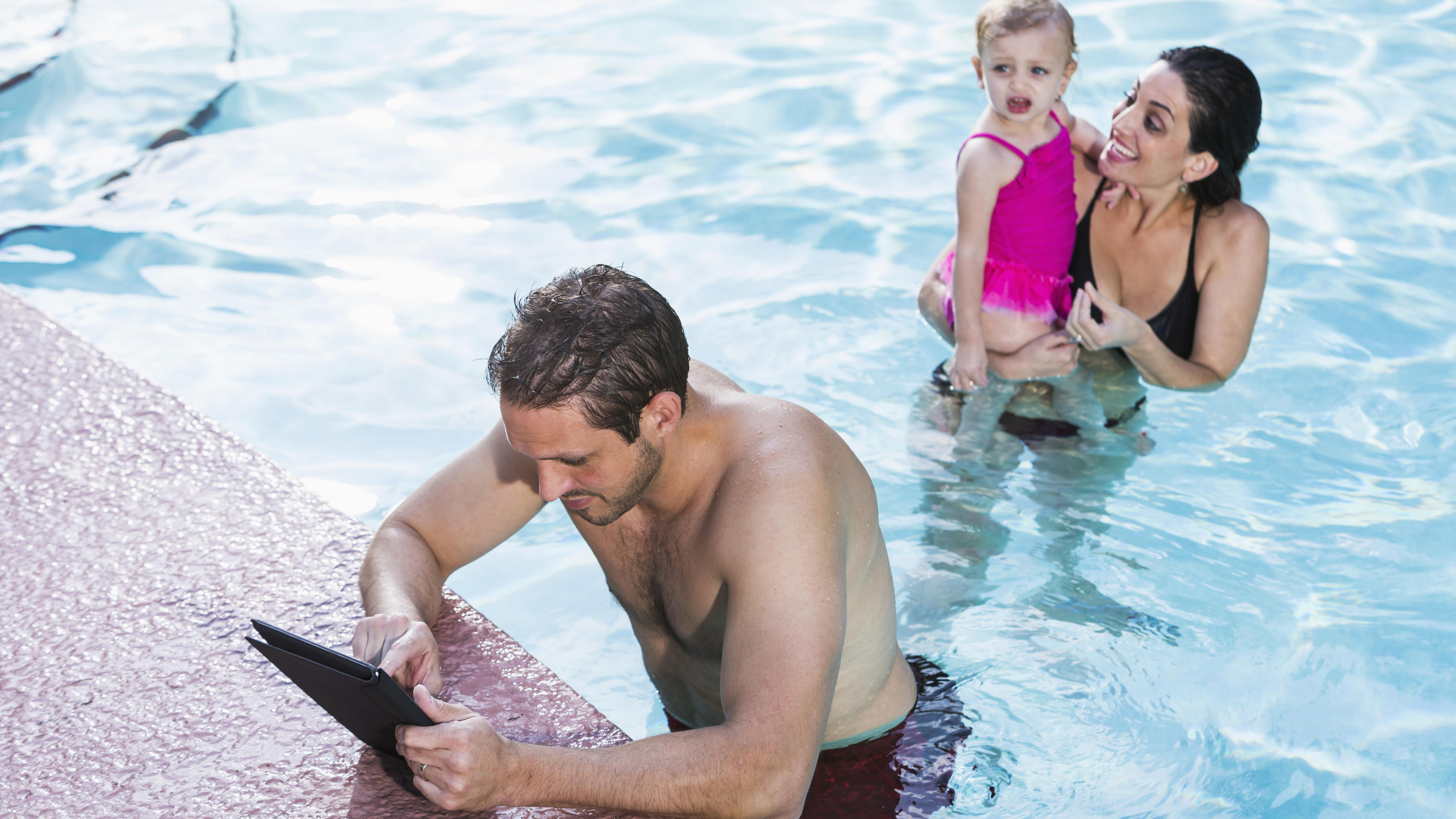 Ein Mann hängt im Schwimmbad am Beckenrand und schaut auf sein iPad, im Hintergrund sieht man eine Frau mit Kind.