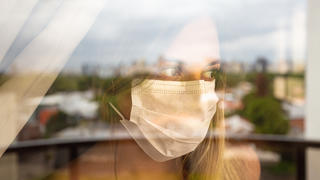 Junge Frau mit Maske steht vor ihrem Fenster