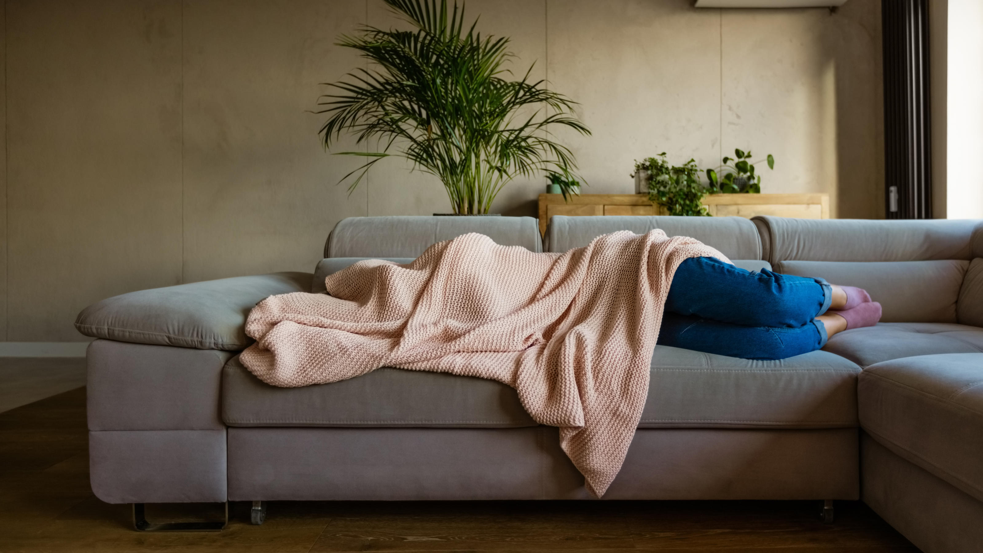 Eine junge Frau liegt auf ihrem Sofa im Wohnzimmer und hat sich die Decke über den Kopf gezogen.