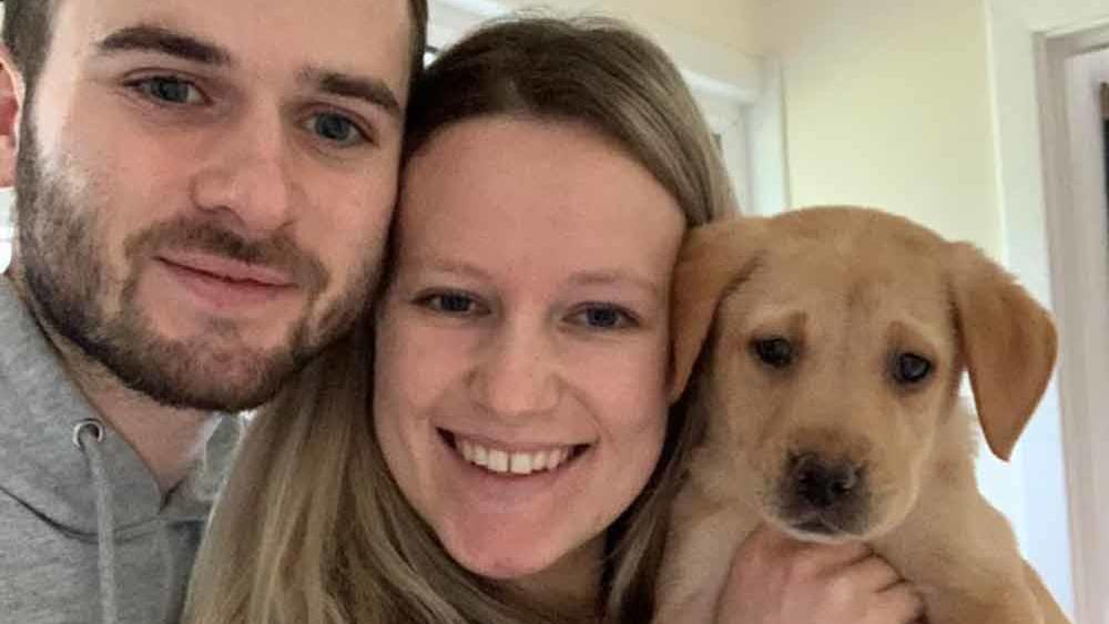 Kate und Ryan halten ihren Labrador-Welpen Coby in die Kamera und lachen.