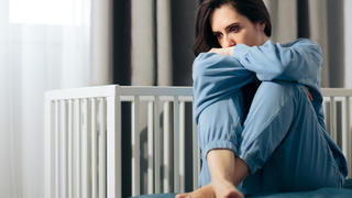 Eine Frau sitzt nachdenklich und traurig aussehend neben einem Babybett.