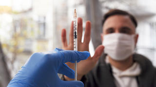 Ein Mann mit Mund-Nasenschutz-Maske steht im Hintergrund, streckt seine Hand in Richtung Kamera aus, um eine Spritze mit einer Impfung abzulehnen.