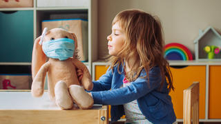 Ein kleines Mädchen hat ihrem Kuscheltier eine Mund-Nasenschutz-Maske aufgesetzt.