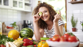 Eine junge, glückliche Frau umgeben von gesundem Essen, wie Obst und Gemüse.