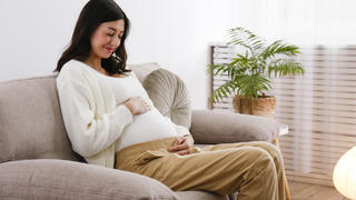 Eine junge Frau sitzt auf ihrem Sofa und hält sich ihren Schwangerschaftsbauch.