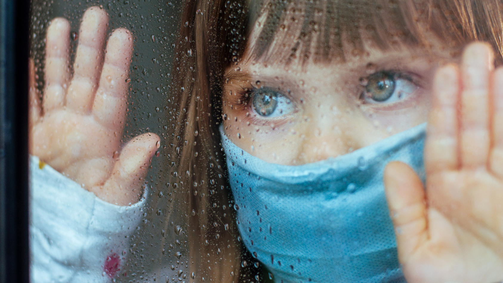 Mädchen steht mit Corona-Maske am Fenster und blickt traurig nach draußen