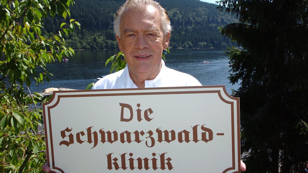 Schauspieler Klausjürgen Wussow hält vor der Kulisse des Titisees im Schwarzwald ein Schild mit der Aufschrift "Die Schwarzwaldklinik" in den Händen