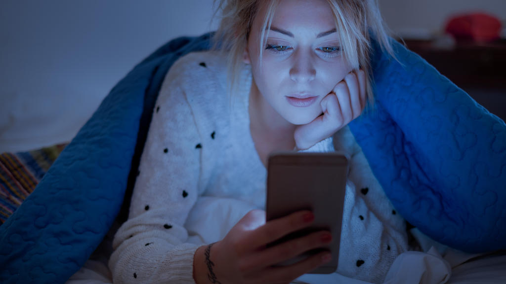 Eine junge Frau liegt im Bett und schaut auf ihr Smartphone. Dabei sieht sie alles andere als glücklich aus.