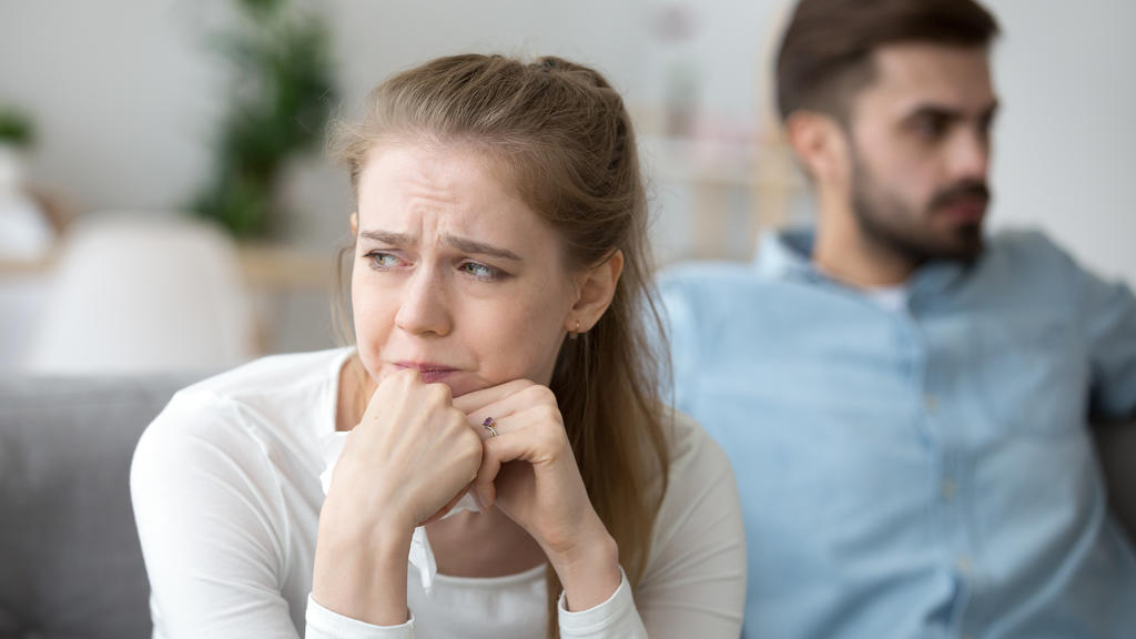 Ein junges Paar sitzt auf ihrem Sofa im Wohnzimmer. Die Frau, die im Vordergrund sitzt und von der Kamera fokussiert wird, weint.