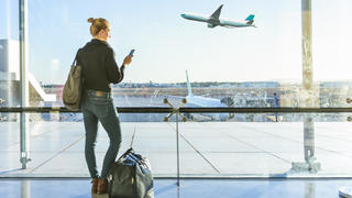Young casual female traveler at airport, holding smart phone device, looking through the airport gate windows at planes on airport runway.