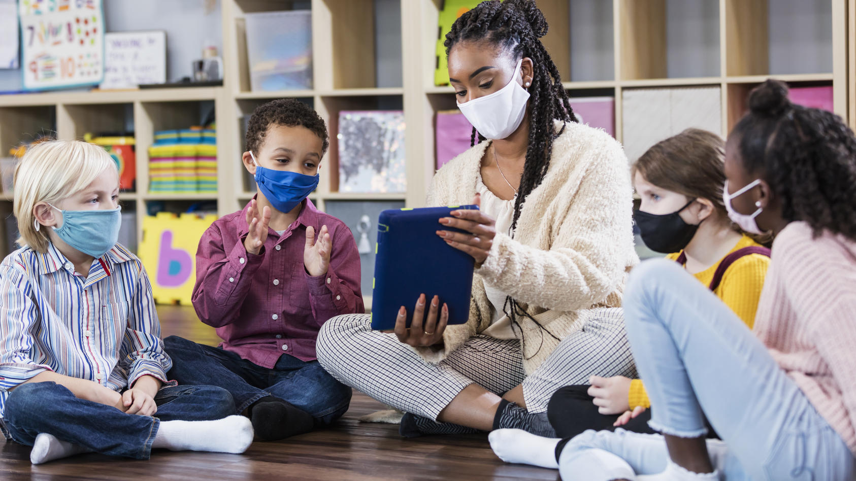 Eine Gruppe von Vorschulkinder sitzt in einem Sitzkreis, gemeinsam mit ihrer Erzieherin, die ihnen etwas auf ihrem Tablet zeigt.
