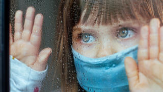 Mädchen steht mit Corona-Maske am Fenster und blickt traurig nach draußen