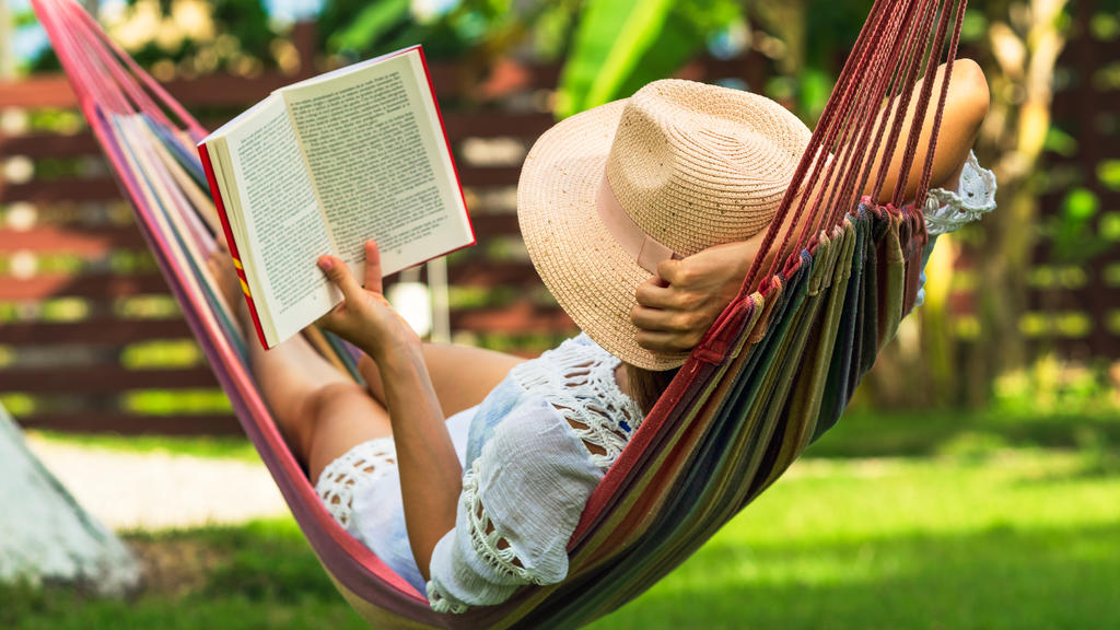 Eine Frau spannt auf einer Hängematte im Garten aus und liest ein Buch.
