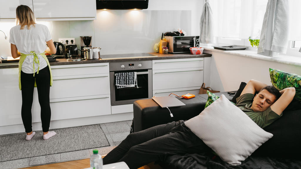 Eine Frau steht in der Küche und kocht während ihr Partner auf dem Sofa liegt, faulenzt und nichts tut.