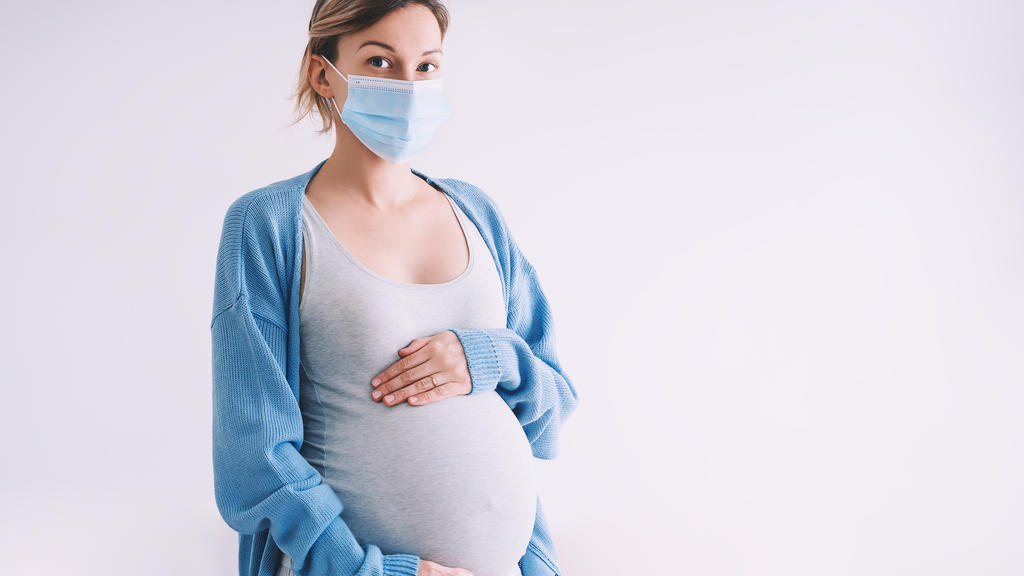 Eine schwangere Frau hält ihren Bauch und trägt eine Mund-Nasenschutz-Maske.