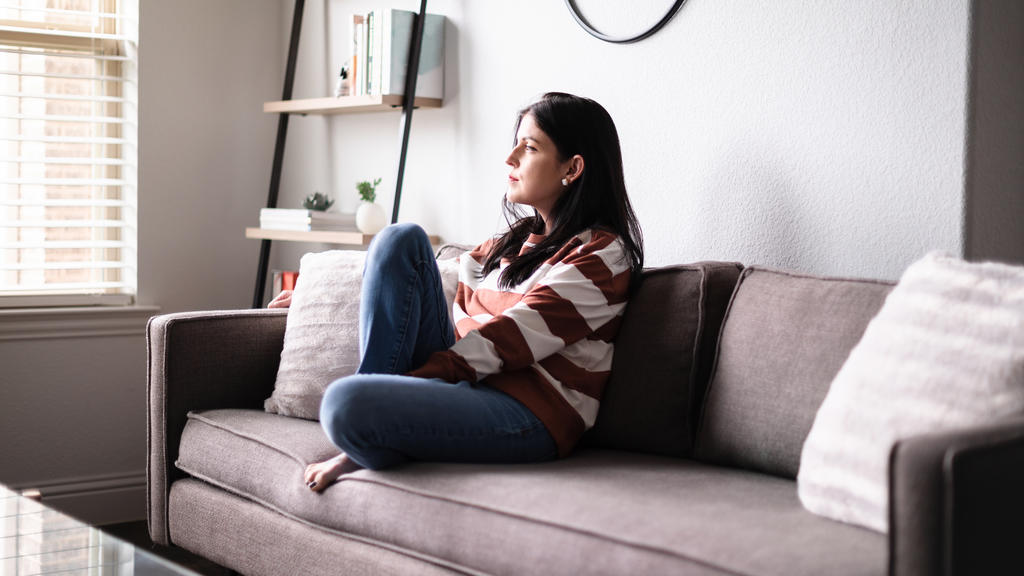 Eine junge Frau sitzt auf ihrem Sofa und schaut nachdenklich in die Luft.