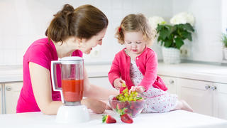 Beautiful young mother and her happy laughing toddler girl making fresh strawberry and other fruit juice for breakfast together in a sunny white kitchen with a window