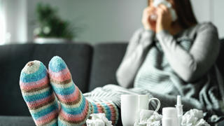 Eine junge Frau sitzt krank auf ihrem Sofa, um sie herum sind unter anderem ganz viele Taschentücher verteilt.