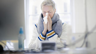 Gestelltes Symbolfoto zum Thema ' krank am Arbeitsplatz '. Eine Frau sitzt sich an ihrem Schreibtisch und putzt sich die Nase.