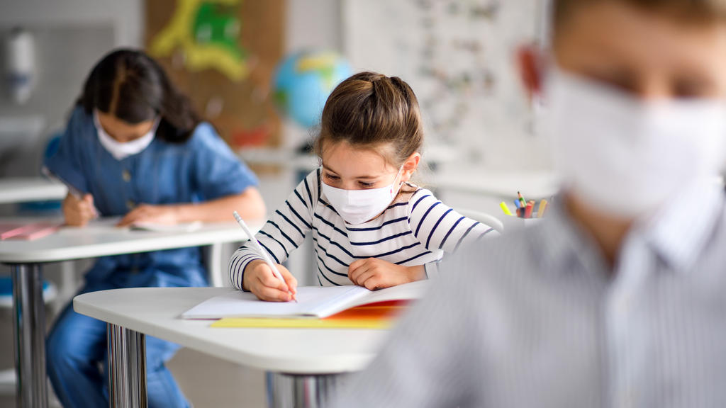Kleinere Kinder sitzen in einem Klassenraum in der Schule, sie tragen Mund-Nasenschutzmaske.