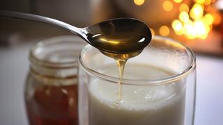 In ein Glas mit warmer Milch wird vor einem beleuchteten Hintergrund ein Löffel Honig gegeben