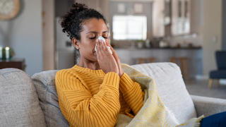 Eine Frau sitzt auf dem Sofa und putzt sich die Nase, weil sie erkältet ist.