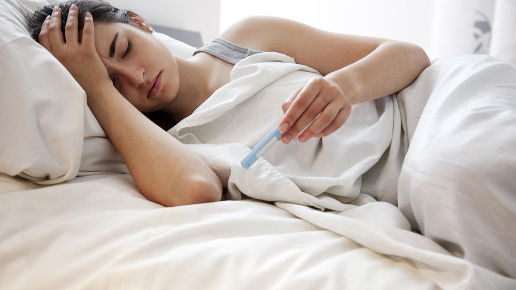 Eine Frau liegt krank, inklusive Fieber-Thermometer, im Bett.