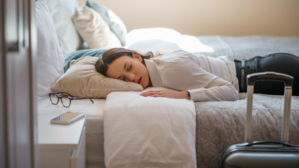 Eine Geschäftsfrau liegt mitsamt ihren Klamotten auf ihrem Hotelbett und scheint völlig erschöpft zu sein.