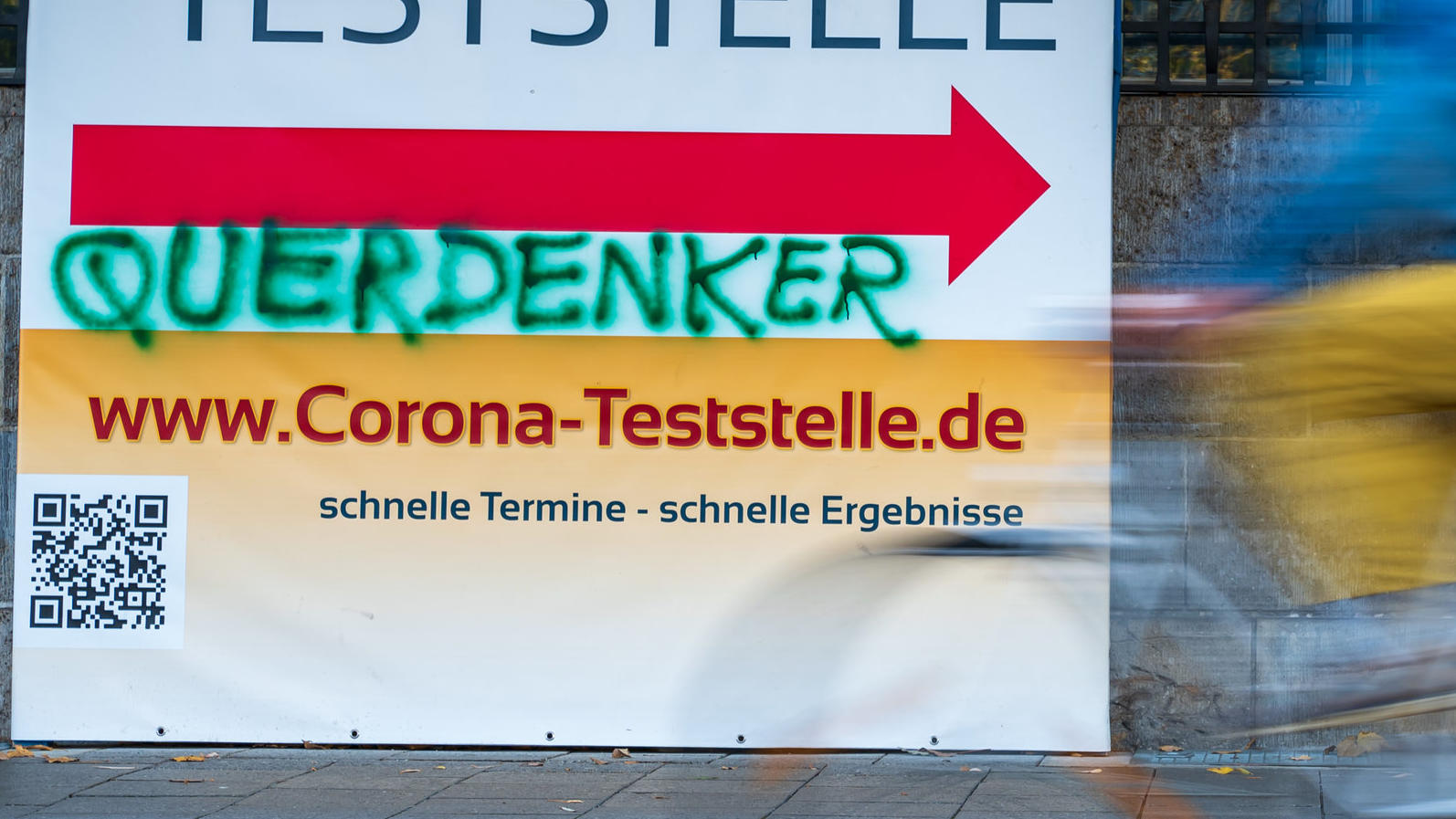 ARCHIV - 02.11.2021, Bayern, München: Ein Mann radelt an einem Plakat vorbei, auf das jemand das Wort «Querdenker» gesprüht hat. «Querdenker» und andere Begriffe im Zusammenhang mit der Corona-Pandemie dominieren auch die Suche nach dem Unwort des Ja