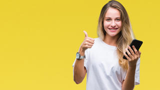 Eine junge blonde Frau steht vor einer gelben Wand und hält ihren Daumen hoch. In der anderen Hand hält sie ihr Smartphone.