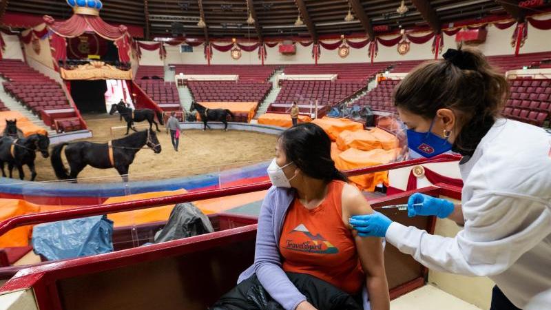 Impfärztin Caroline Klima (r) impft im Circus Krone Paitul Supaporn gegen das Corona-Virus, während im Hintergrund Pferde in der Manege trainieren. Foto: Lennart Preiss/dpa