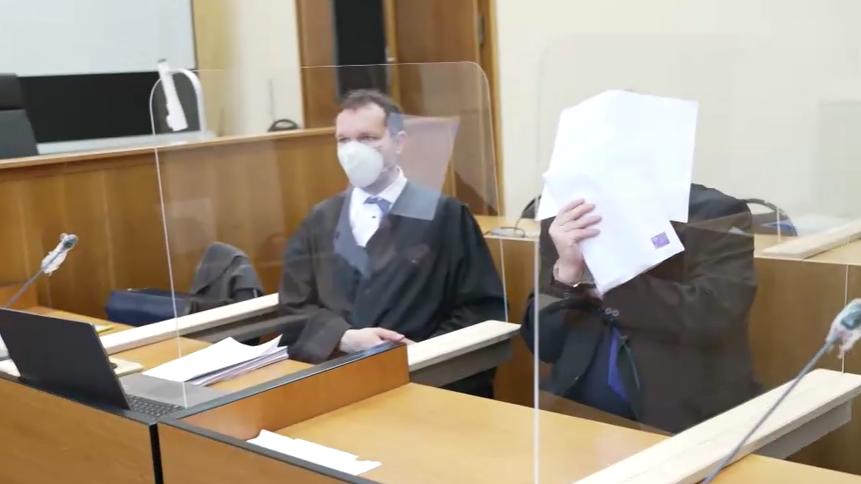 Zwei Männer im Gerichtssaal, einer hält sich Papier vor das Gesicht.