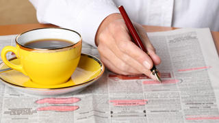 Eine junge Frau streicht sich Stellenanzeigen in der Zeitung an, daneben steht eine Tasse Kaffee.