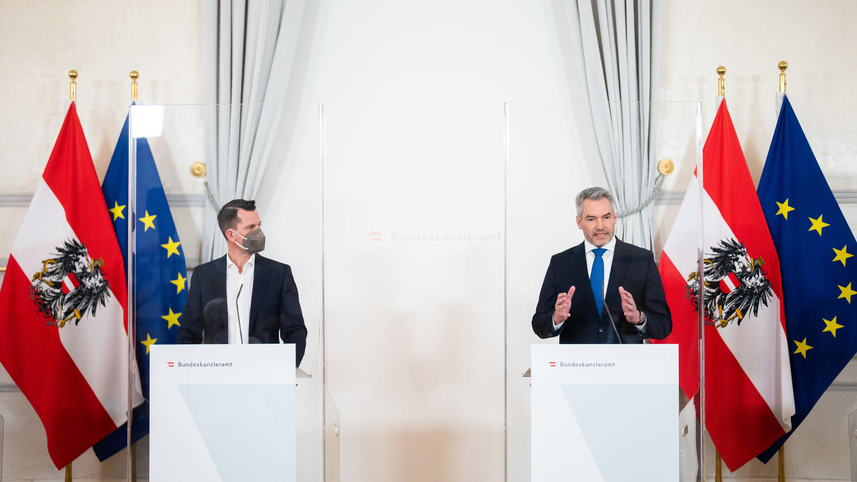 Gesundheitsminister Wolfgang Mückstein (Grüne) und der österreichische Bundeskanzler Karl Nehammer (ÖVP) stehen an zwei weißen Rednerpulten. Im Hintergrund hängen zwei Österreich- und zwei EU-Flaggen. 