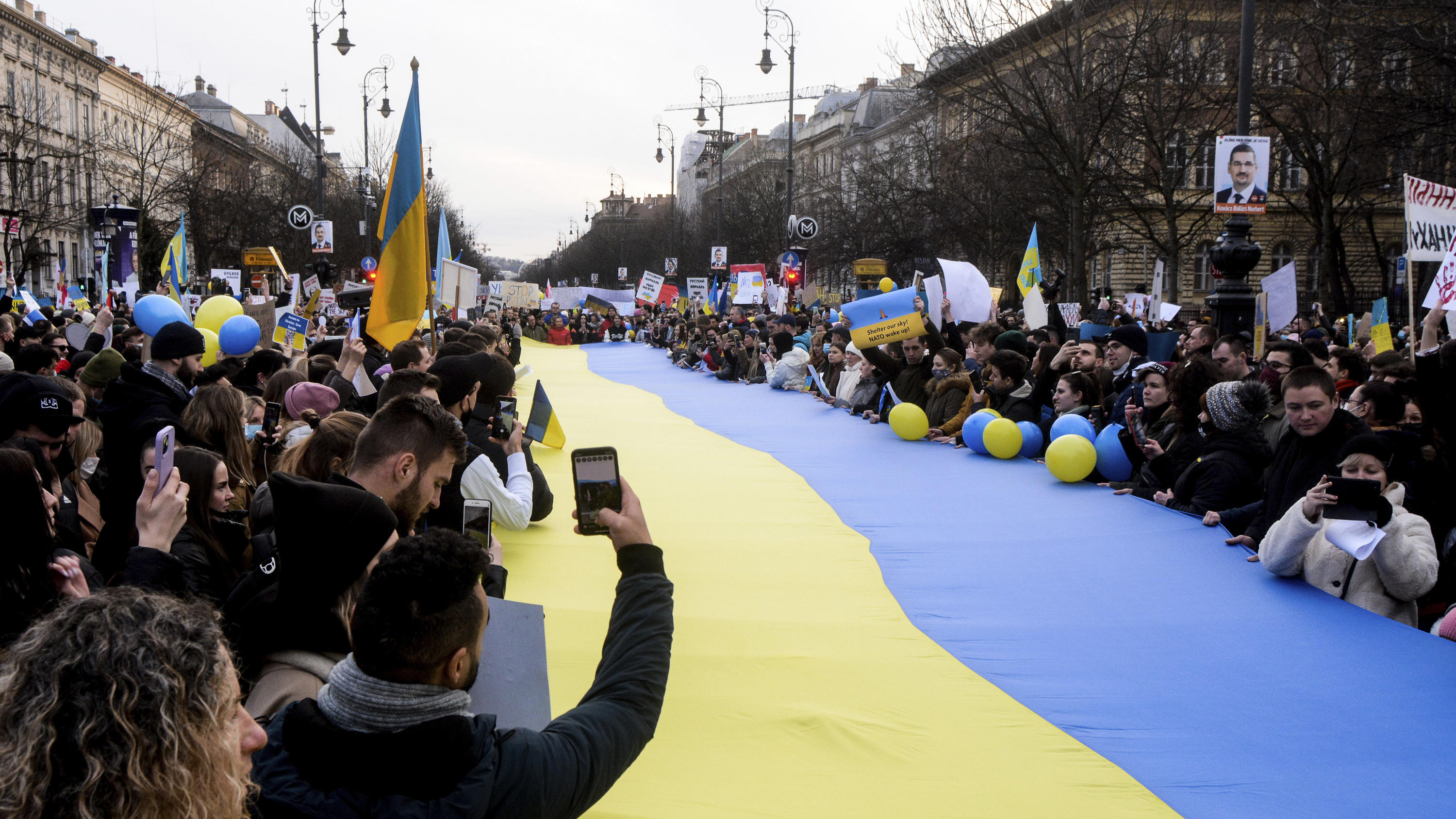 26.02.2022, Ungarn, Budapest: Demonstranten stehen um eine große ukrainische Flagge versammelt und protestieren gegen den russischen Einmarsch in die Ukraine. Russische Truppen haben den erwarteten Angriff auf die Ukraine gestartet und drangen in die