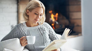 Eine junge Frau sitzt mit einer Tasse Tee und einem Buch vor einem Kamin und lässt es sich gut gehen.