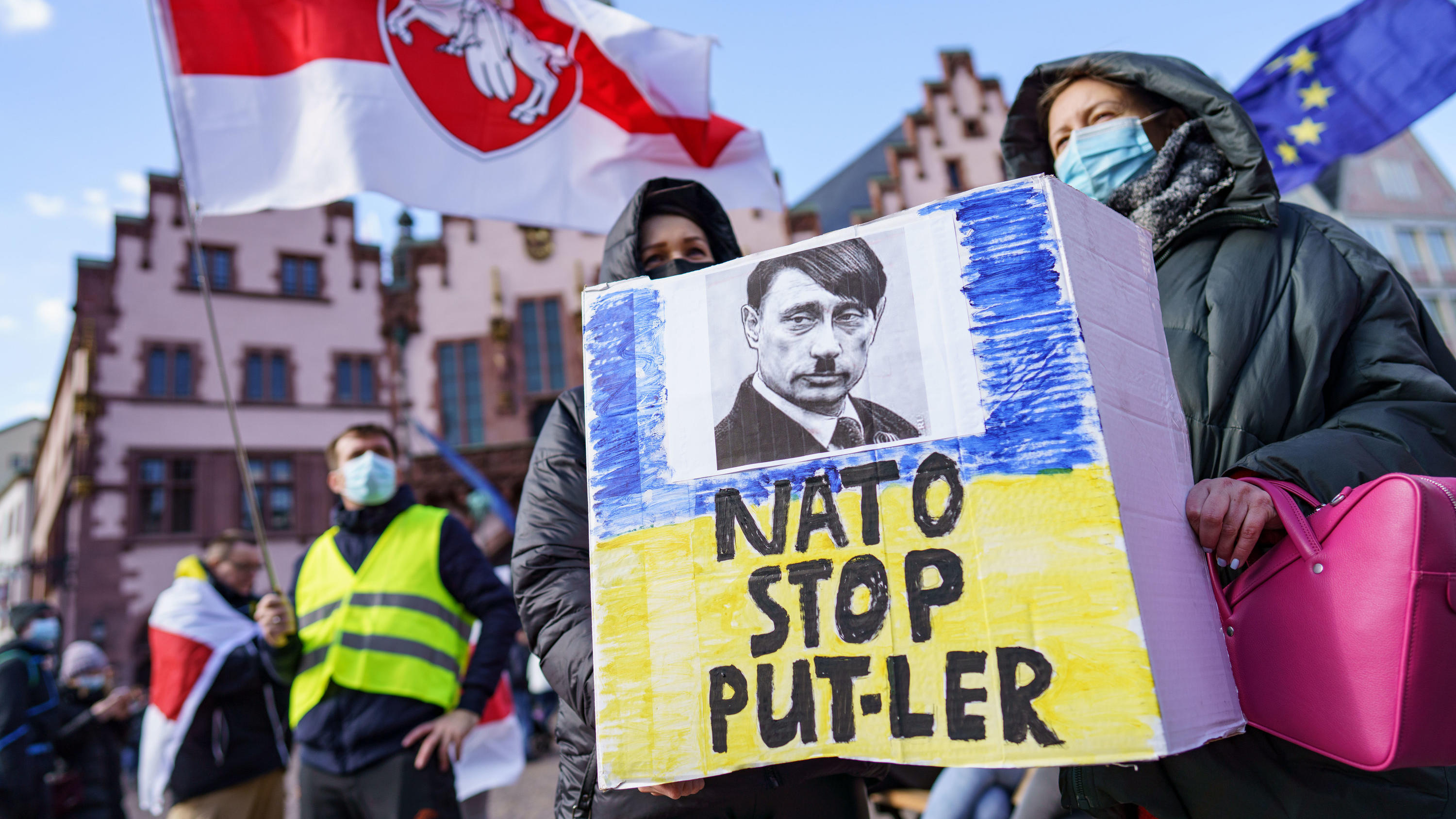 27.02.2022, Hessen, Frankfurt/Main: Demonstrantinnen halten ein Plakat mit der Aufschrift "Nato stop Put-ler" mit dem Bild eines Gesichts, das eine Mischung von Hitler und Putin darstellt, bei einer Kundgebung auf dem Römerberg für Frieden in der Ukr