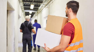 Arbeiter mit Warnweste trägt Kartons durch Gang zum Lager einer Fabrik || Modellfreigabe vorhanden