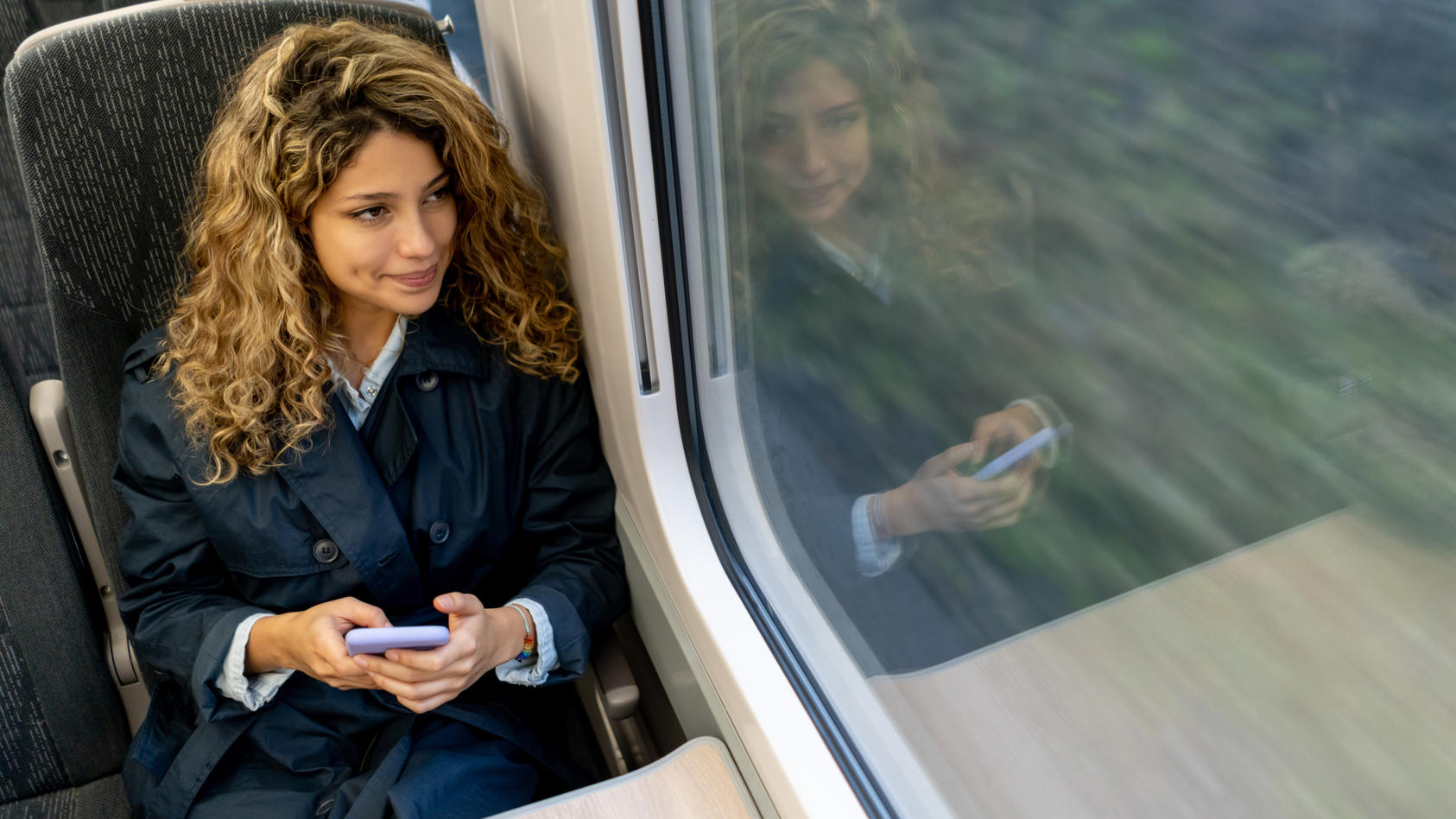 Eine Frau sitzt im Zug und schaut aus dem Fenster. In ihrer Hand hält sie ihr Handy.