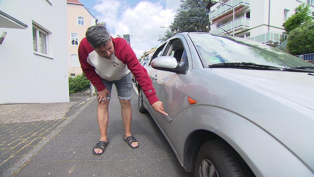 Ein Mann bückt sich und zeigt auf die Kratzer auf einem Auto in Wiesbaden.
