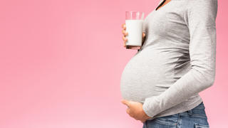 Eine schwangere Frau liegt auf dem Sofa, streichelt ihren Bauch und hält dabei ein Glas Milch in der Hand.
