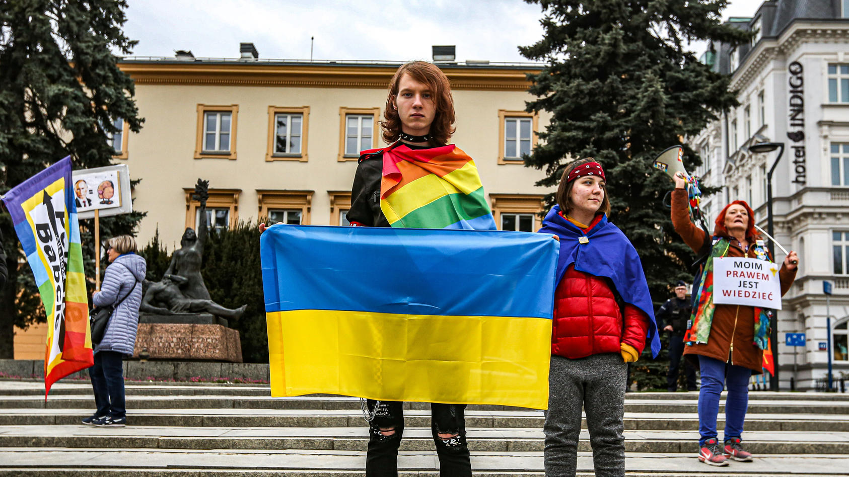 Ein junger Aktivist im Vordergrund mit einem ernsten Gesichtsausdruck, einer Ukraineflagge in der Hand und einer Regenbogenflagge um den Körper gewickelt.